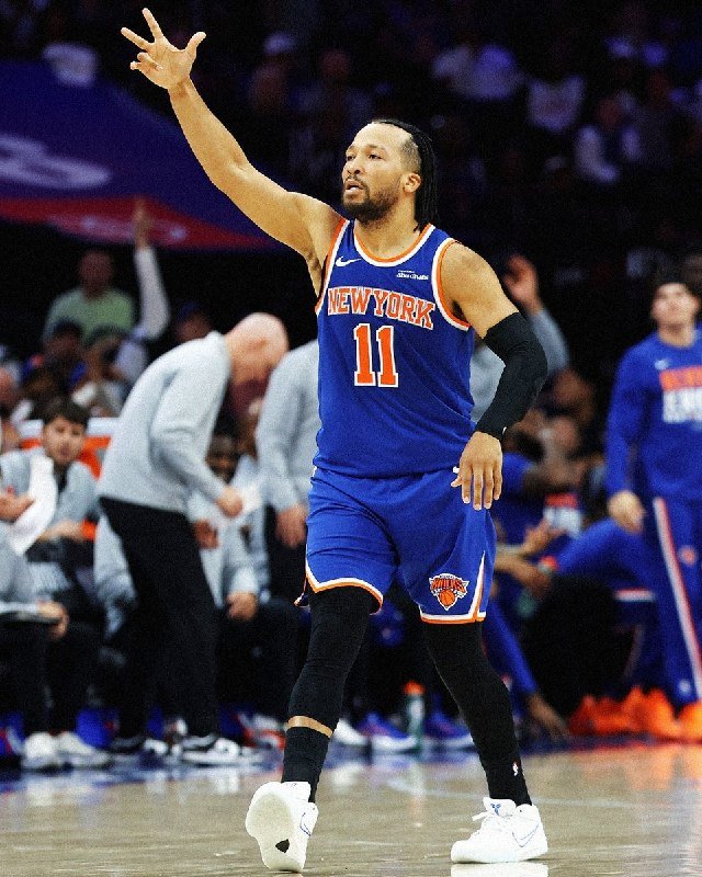 A basketball player wearing a blue "NEW YORK" jersey with the number 11 raises his arm with three fingers extended on a court.