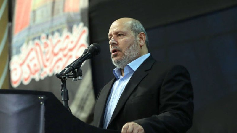 A bearded man in a dark suit and blue shirt speaks at a podium into a microphone, with a banner featuring Arabic script visible in the background.