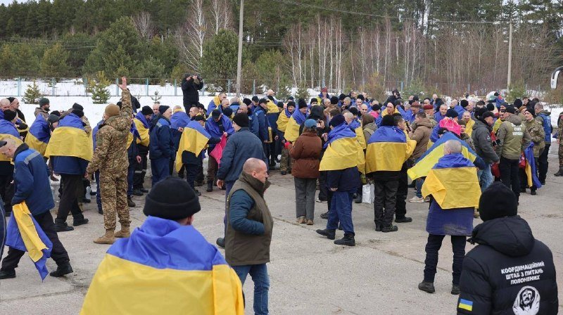 A crowd of people draped in blue and yellow flags gathers outdoors in a snowy area with bare trees in the background.