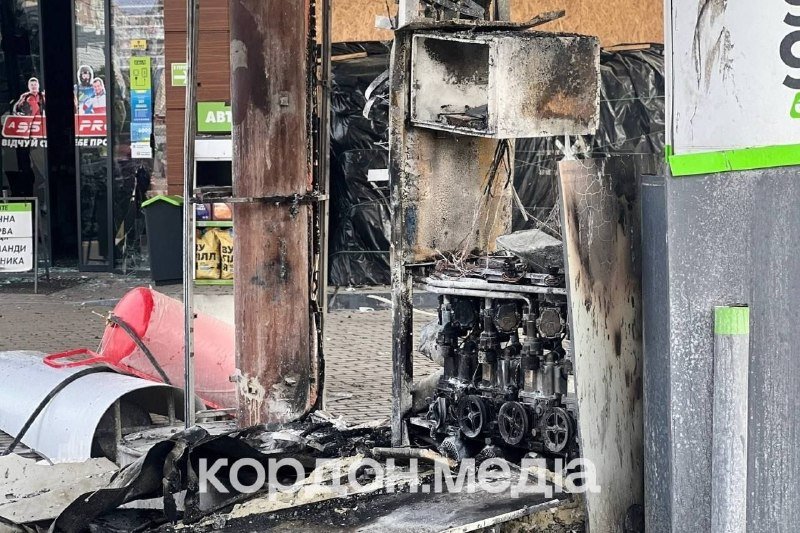 A blackened, charred ATM machine sits gutted on a street, its metal panels melted and burnt debris scattered across the pavement in front of storefronts.