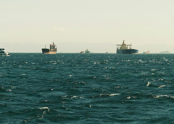 Several cargo ships are visible on a choppy, dark blue sea under a hazy sky, with faint landforms appearing on the distant horizon.