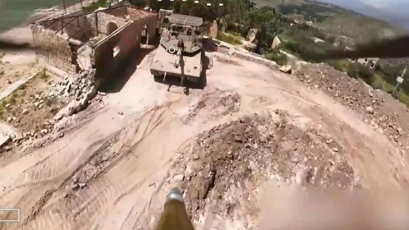 An armored military vehicle is parked on a dirt clearing beside a stone structure, viewed from an elevated camera perspective.