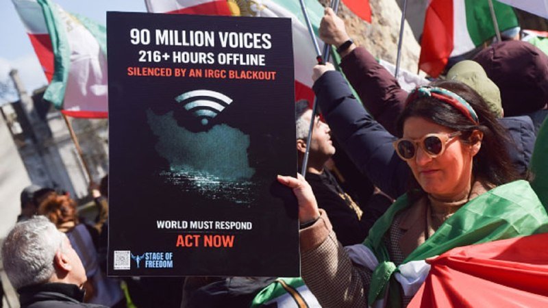 A woman at a protest holds a sign reading "90 MILLION VOICES, 216+ HOURS OFFLINE, SILENCED BY AN IRGC BLACKOUT" alongside others waving Iranian flags.