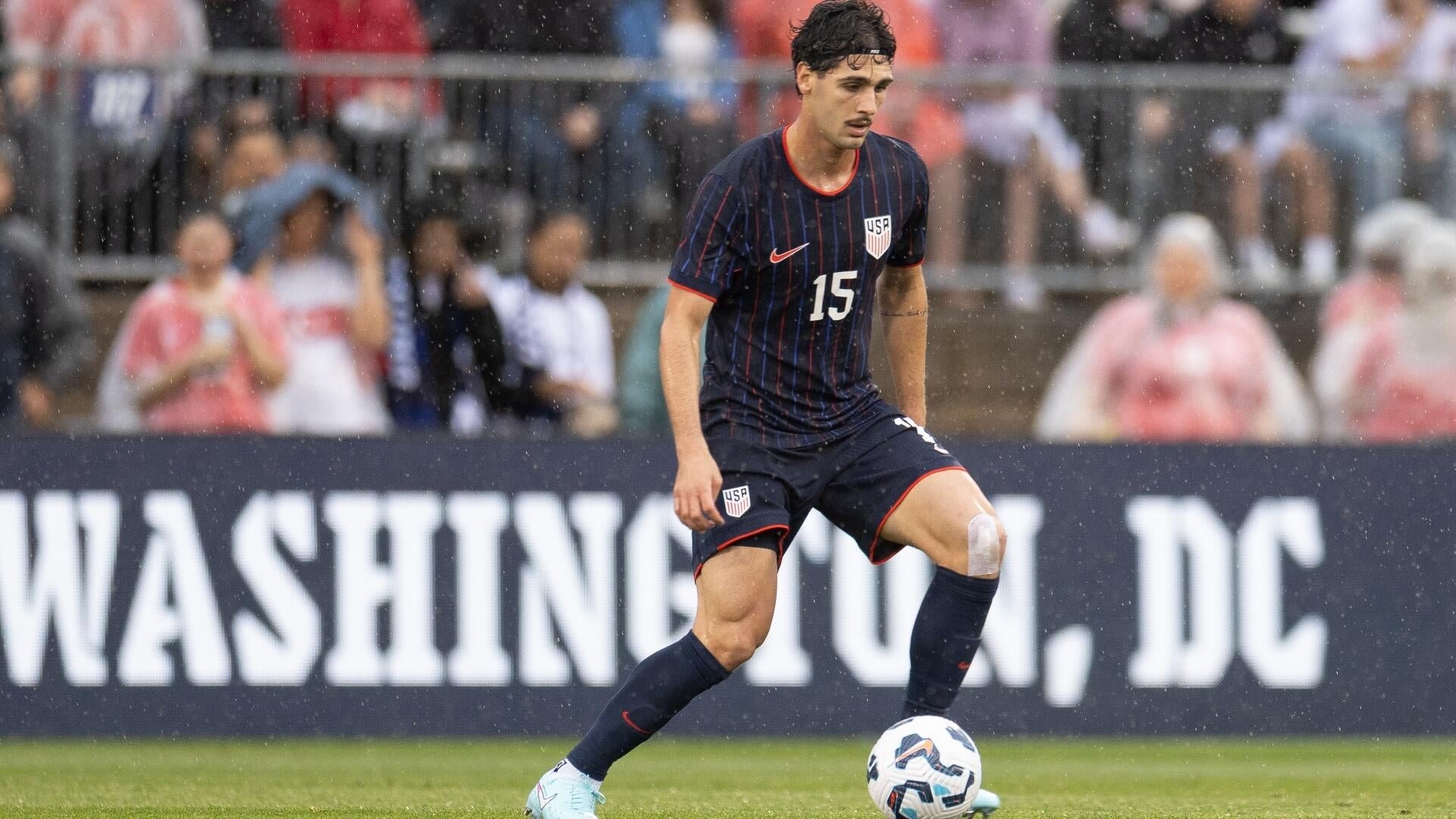A soccer player wearing a navy USA jersey with the number 15 controls a ball on the field in rainy conditions, with a "WASHINGTON, D.C." banner visible behind him.