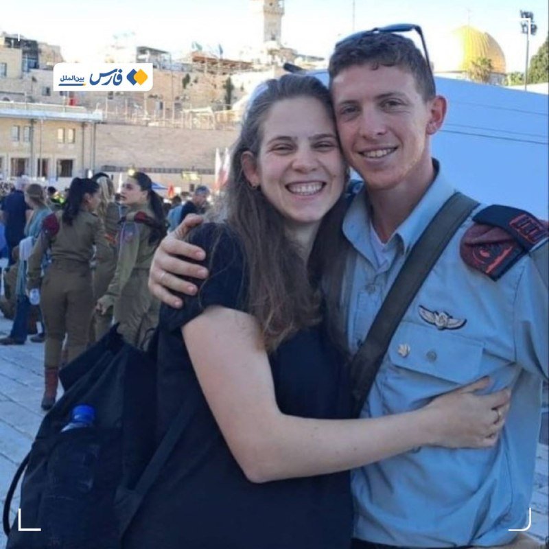 A smiling woman in a black top embraces a man in a light blue military uniform with insignia, standing before a stone wall and a golden-domed building.