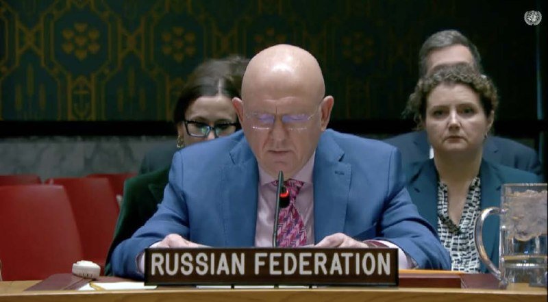 A bald man in a blue suit and glasses speaks at a desk bearing a "RUSSIAN FEDERATION" placard, with other attendees seated behind him.
