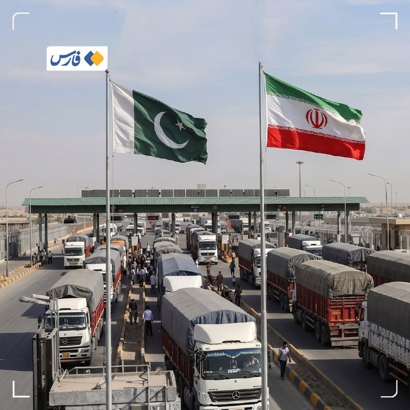 Trucks lined up at a border checkpoint beneath two large flags, one green and white with a crescent, the other green, white, and red with an emblem.