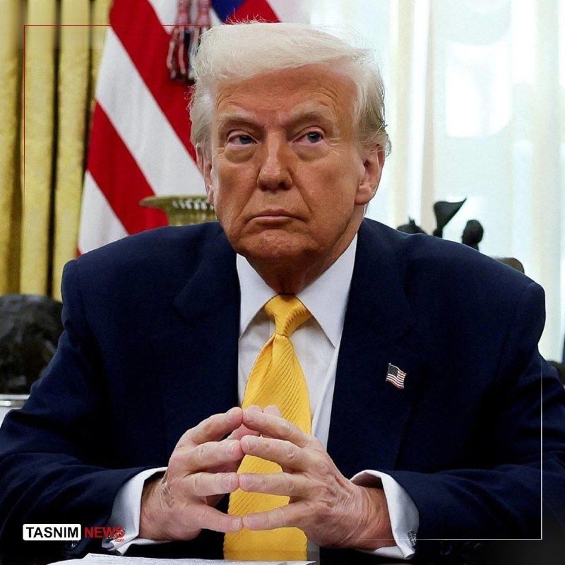 An older man with blonde hair in a dark suit and yellow tie sits with hands clasped, with an American flag visible behind him.