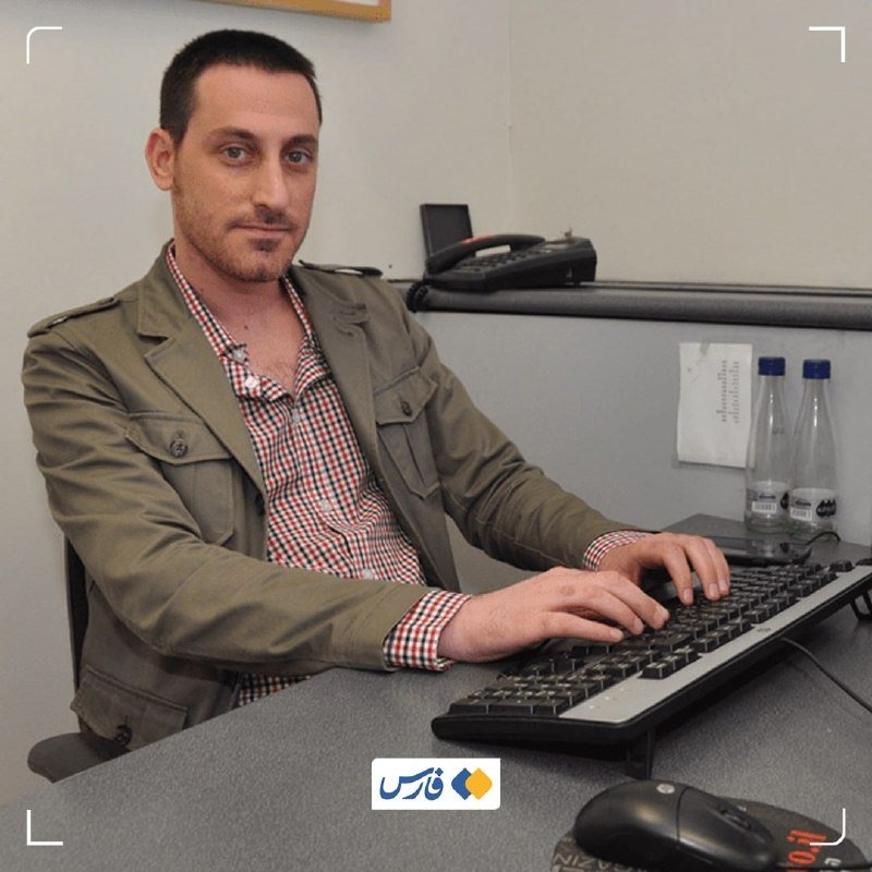 A man in an olive jacket and checkered shirt sits at a desk typing on a keyboard, with a telephone, water bottles, and a computer mouse visible.