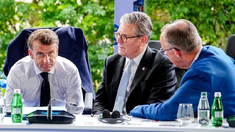 Three men in suits sit at a conference table with microphones, water bottles, and a nameplate, appearing to engage in a formal discussion in a room with greenery visible behind them.