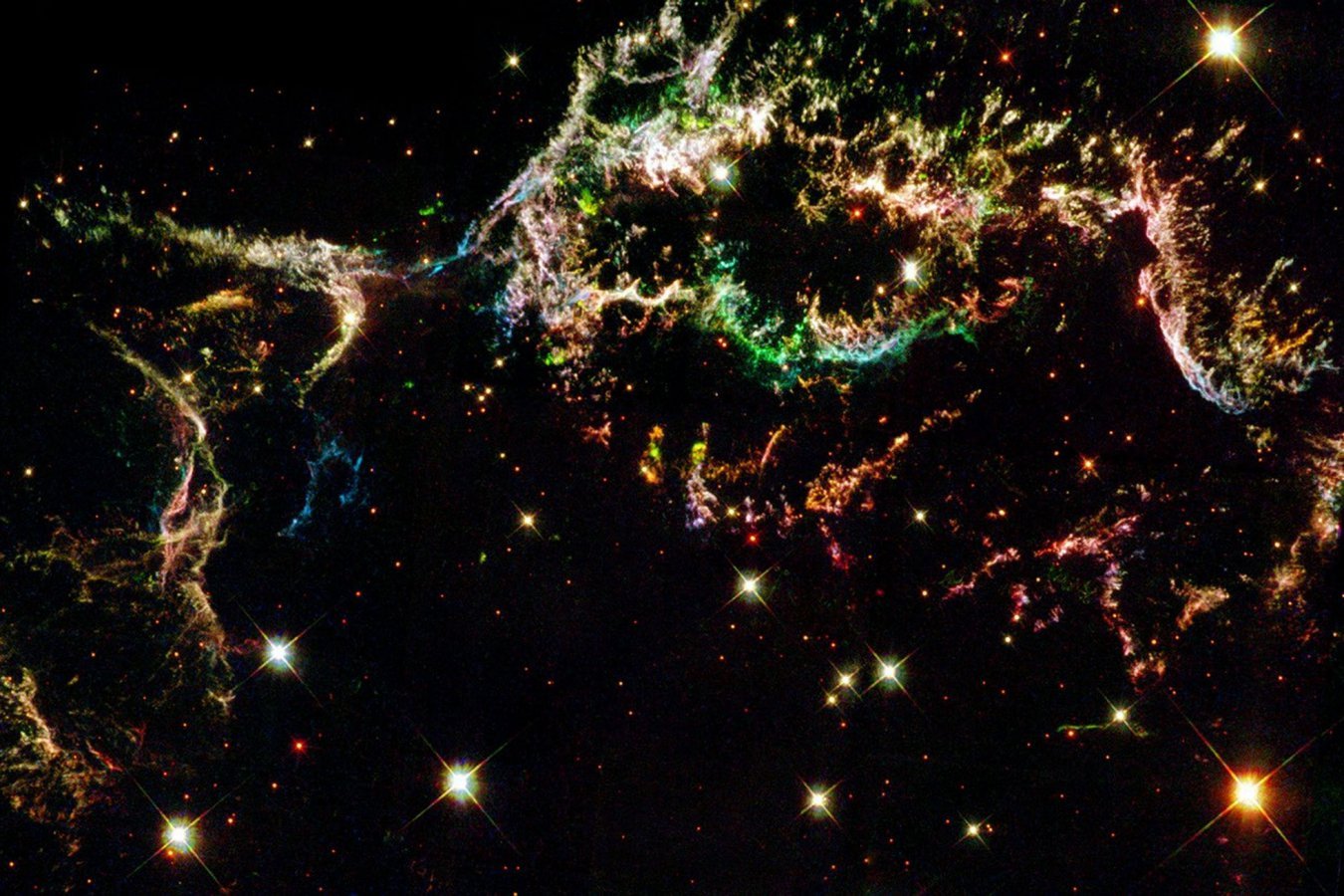 Colorful wispy filamentary structures of a nebula glow in green, pink, orange, and blue against a black background dotted with bright stars.