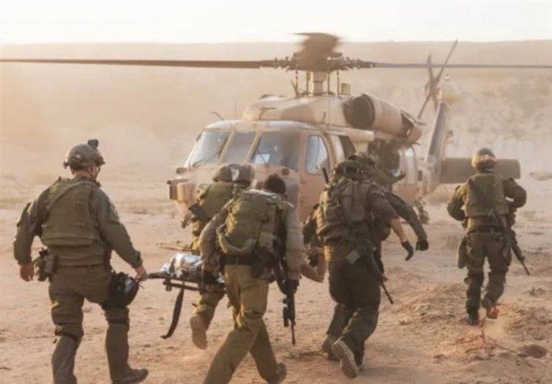 Armed soldiers carry a wounded comrade on a stretcher toward an idling helicopter in a dusty, arid landscape.