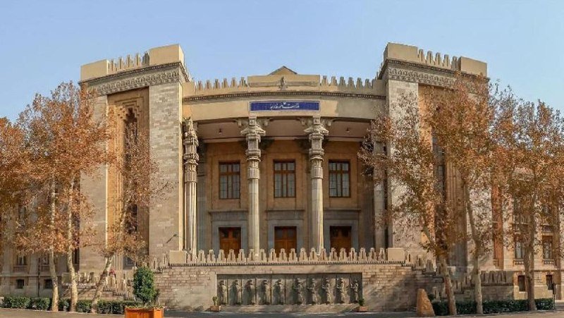 A grand stone building with columns, crenellated towers, and Arabic script on its facade stands behind rows of statues and autumn trees.