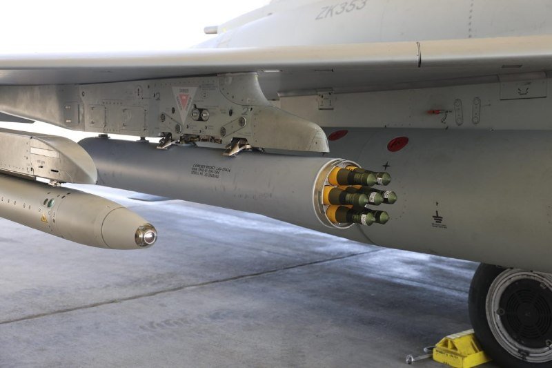 A military jet aircraft parked on a tarmac displays a rotary cannon pod with multiple green-tipped barrels and an attached munition beneath its wing.