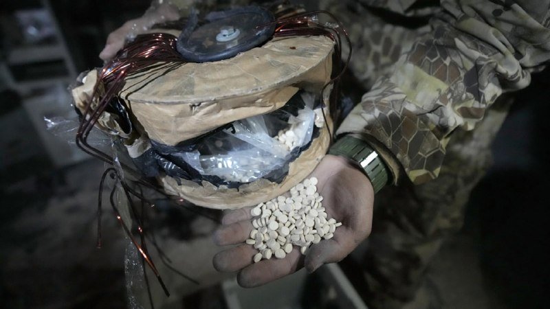 A person in camouflage holds white pills in their palm beside a taped, wire-wrapped cylindrical device containing white powder.