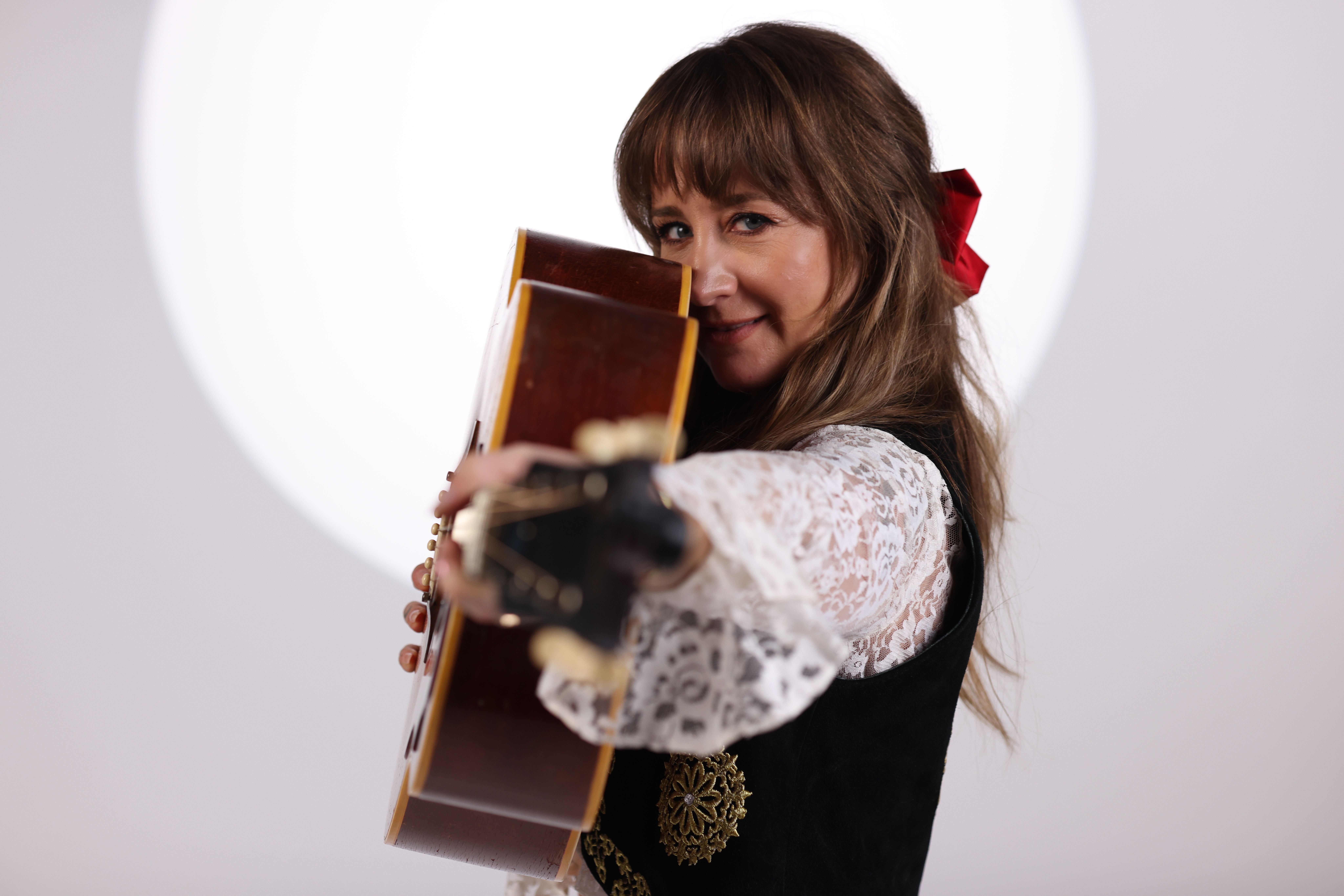 A woman with long brown hair and a red hair bow, wearing a black and white lace blouse, holds an acoustic guitar against a white backdrop.