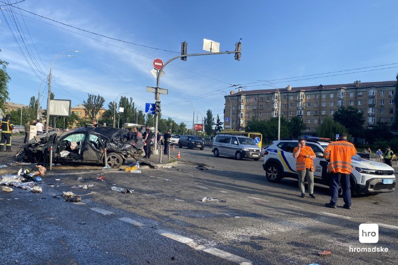 A heavily damaged black car sits on a city intersection as police officers in orange vests investigate, with debris scattered across the road and a police SUV parked nearby.