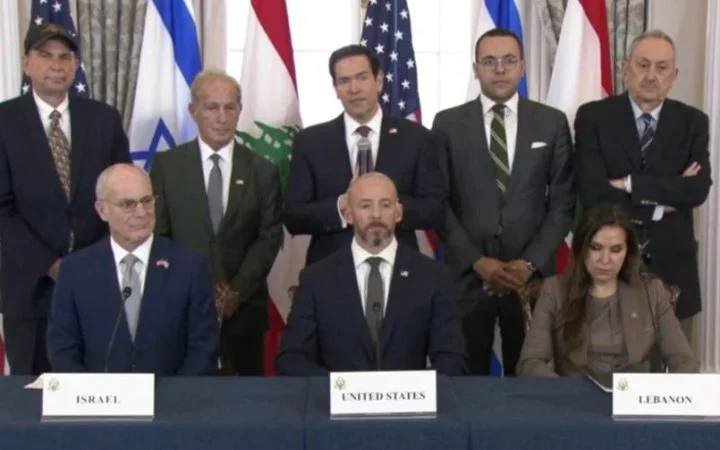 Eight formally dressed individuals pose behind a table with placards reading "ISRAEL," "UNITED STATES," and "LEBANON," with American, Israeli, and Lebanese flags displayed in the background.