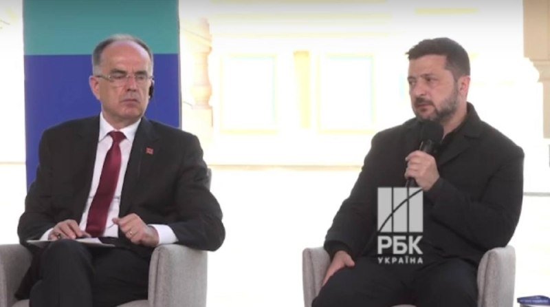 Two men in dark suits sit in armchairs on a stage with a teal and blue backdrop, one wearing a Turkish flag pin and the other speaking into a microphone.