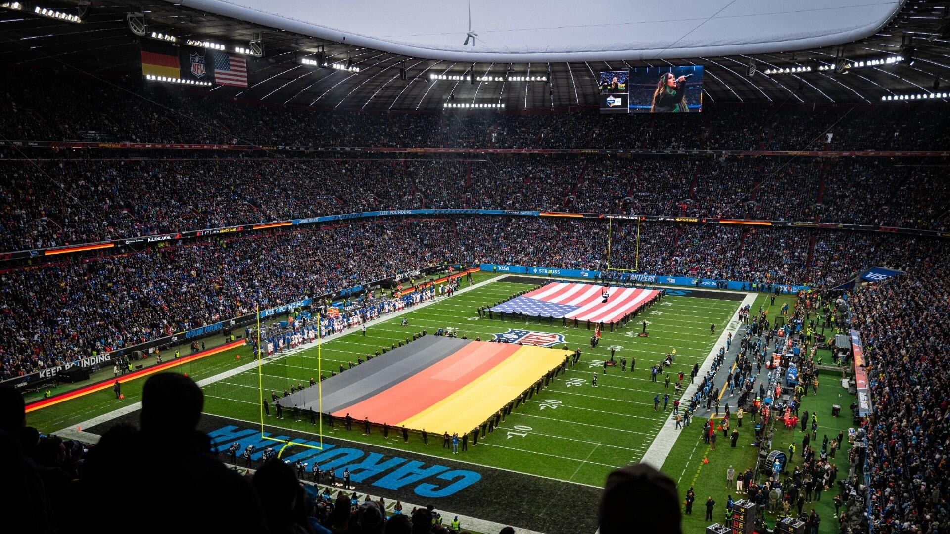A large American flag and a German flag are unfurled on the field of a packed NFL stadium during a pre-game ceremony, with players and staff lined up along the sidelines.