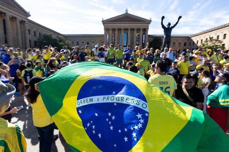 A large crowd wearing yellow and green Brazilian jerseys gathers in a plaza, with a giant Brazilian flag held in the foreground and a bronze statue visible behind them.