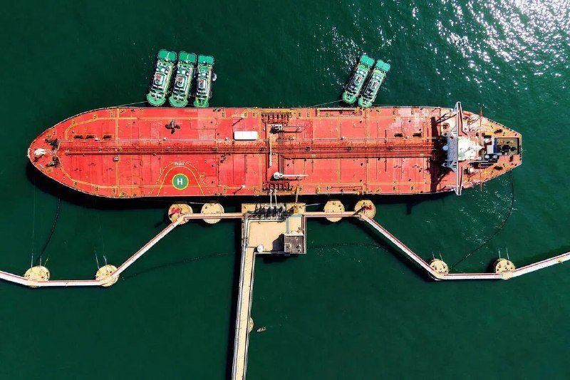 Aerial view of a large red oil tanker marked with an "H" docked at a pier, accompanied by five small green boats in green water.