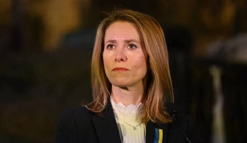 A woman with long blonde hair wearing a dark blazer and white top stands against a dark background, with a blue and yellow ribbon visible on her lapel.