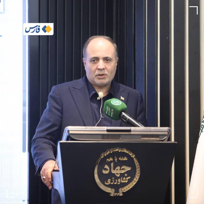 A man in a dark suit stands at a podium with a microphone, with a dark curtain backdrop and an Iranian flag partially visible to the right.