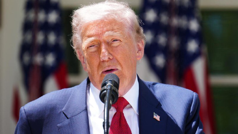 A white-haired man in a blue suit and red tie speaks into a microphone, with two American flags visible in the background.
