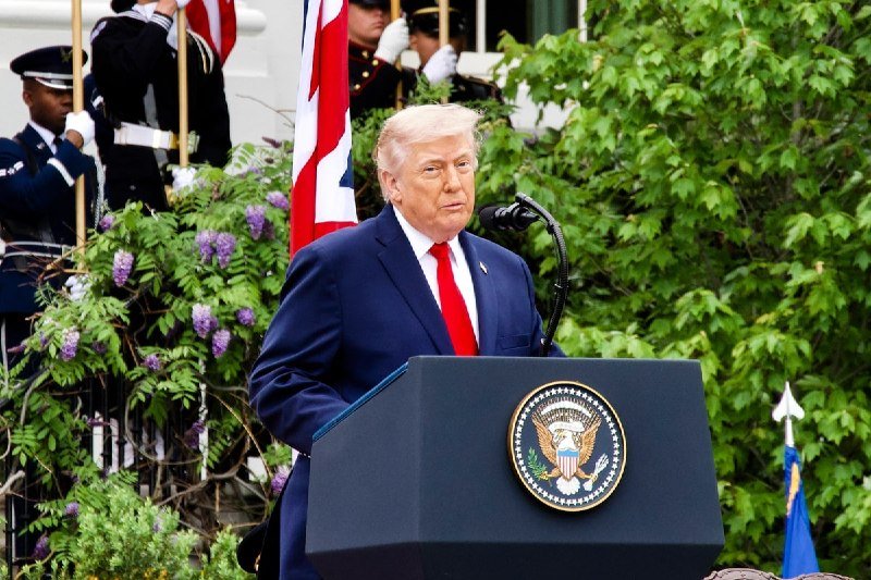 A blond man in a dark blue suit and red tie speaks at a podium bearing the U.S. presidential seal, with military personnel holding flags visible behind green foliage.