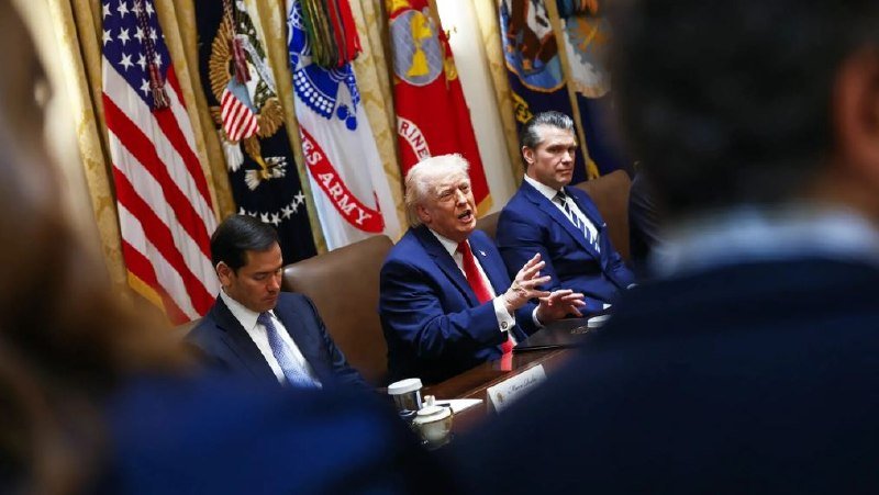 A man in a blue suit and red tie gestures while speaking during a meeting, flanked by two other men in suits, with U.S. and military flags displayed behind them.