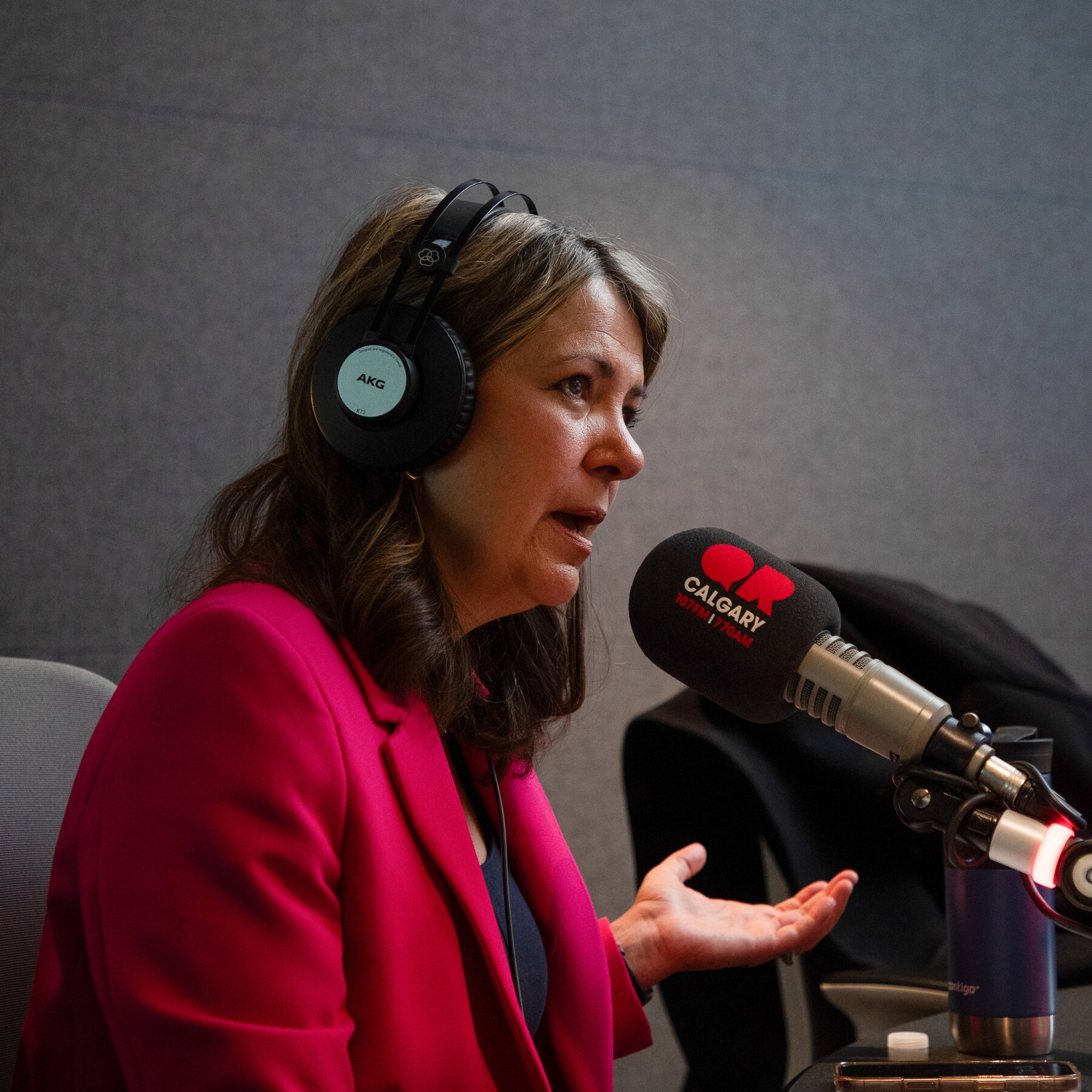A woman in a pink blazer wearing AKG headphones speaks into a microphone labeled "Calgary 107.7 FM" while gesturing with her hand.