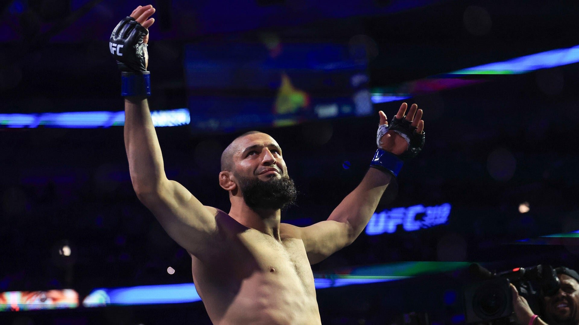 A shirtless bearded man in blue UFC gloves raises both arms upward inside a dimly lit arena.
