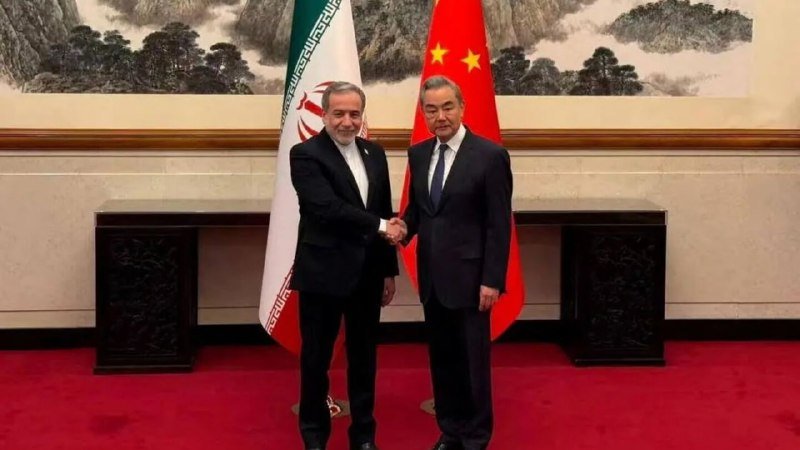 Two men in dark suits shake hands in front of the Iranian and Chinese national flags, with a traditional Chinese landscape painting displayed on the wall behind them.