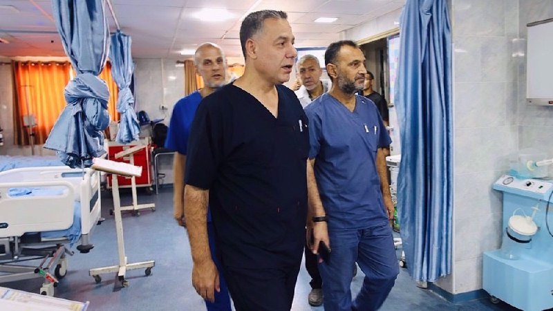 A group of men in medical scrubs walk through a hospital room containing a patient bed and medical equipment.