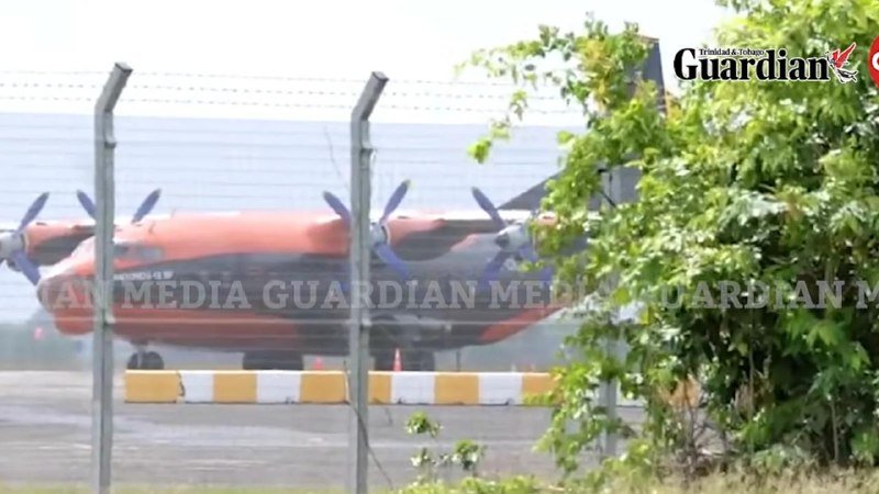 An orange and blue propeller aircraft is parked on a tarmac behind a barbed-wire fence, partially obscured by green foliage.