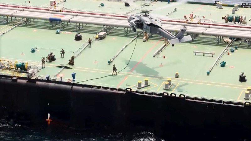 A gray helicopter lands on the green deck of a large vessel as workers in hard hats and military uniforms observe nearby.