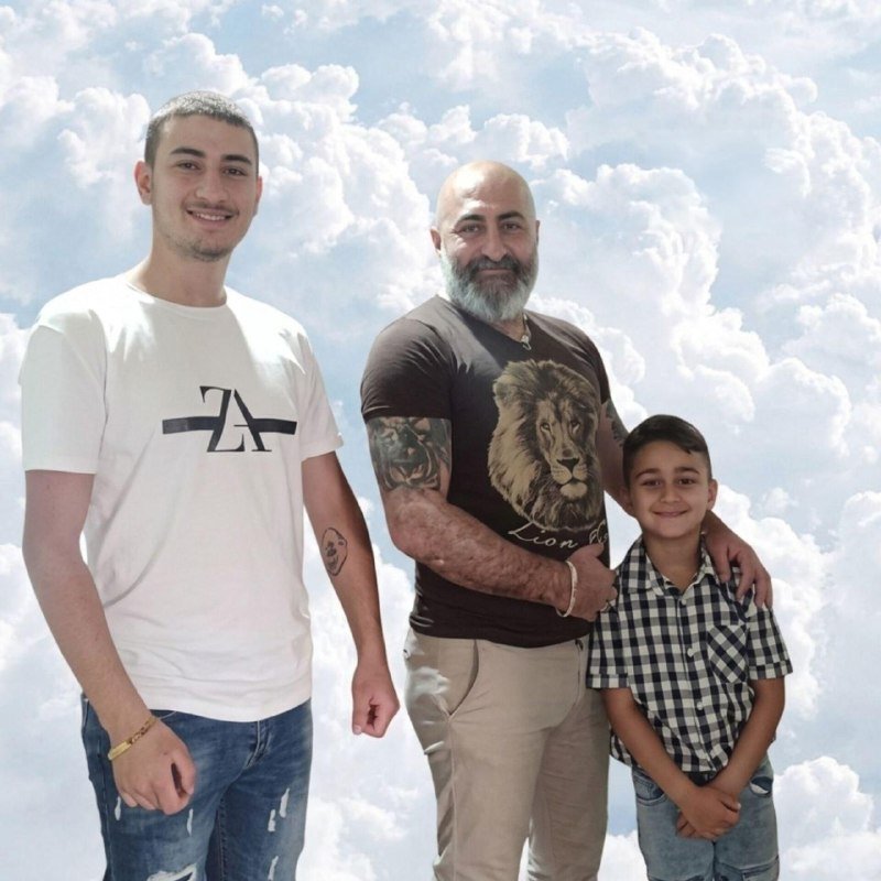Three males pose together against a backdrop of clouds and blue sky, with the central bearded man in a lion-print shirt flanked by a young man in a white t-shirt and a boy in a checkered shirt.