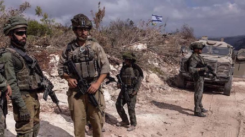 Armed soldiers in green uniforms and helmets stand on a dirt road near a military armored vehicle, with an Israeli flag visible on a distant hillside.