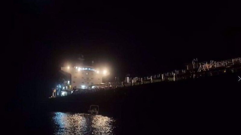 A large illuminated ship is docked at a port at night, with its lights reflecting on the water in the foreground.