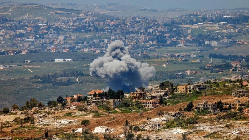 A large plume of gray smoke rises above a hillside village, with a densely built town visible in the distant background.