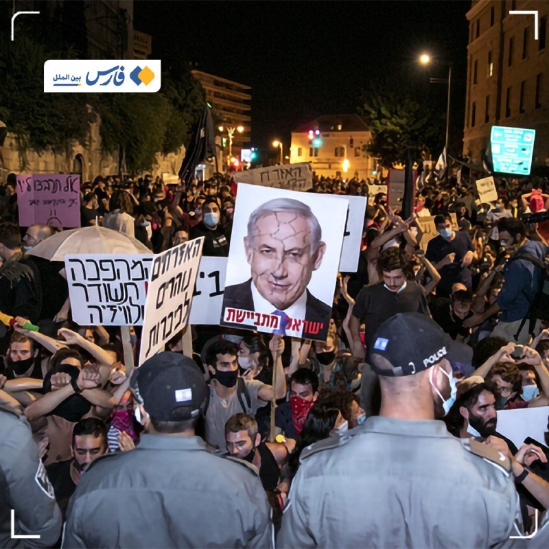Masked protesters hold signs in Hebrew and a portrait placard during a nighttime street demonstration facing police officers in uniform.