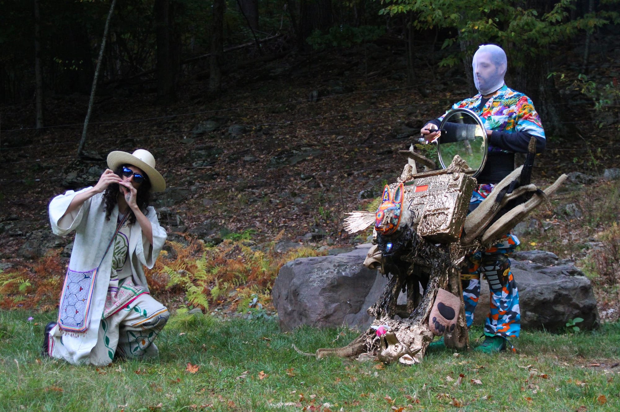 Two masked costumed performers interact in a wooded outdoor setting, one kneeling in white robes and a sun hat with a mirrored sculpture, the other standing in a colorful patterned suit holding a round mirror.