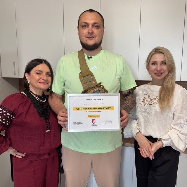 A man in a green t-shirt stands between two women, holding a framed certificate written in Ukrainian that reads "СЕРТИФІКАТ НА КВАРТИРУ" (apartment certificate).