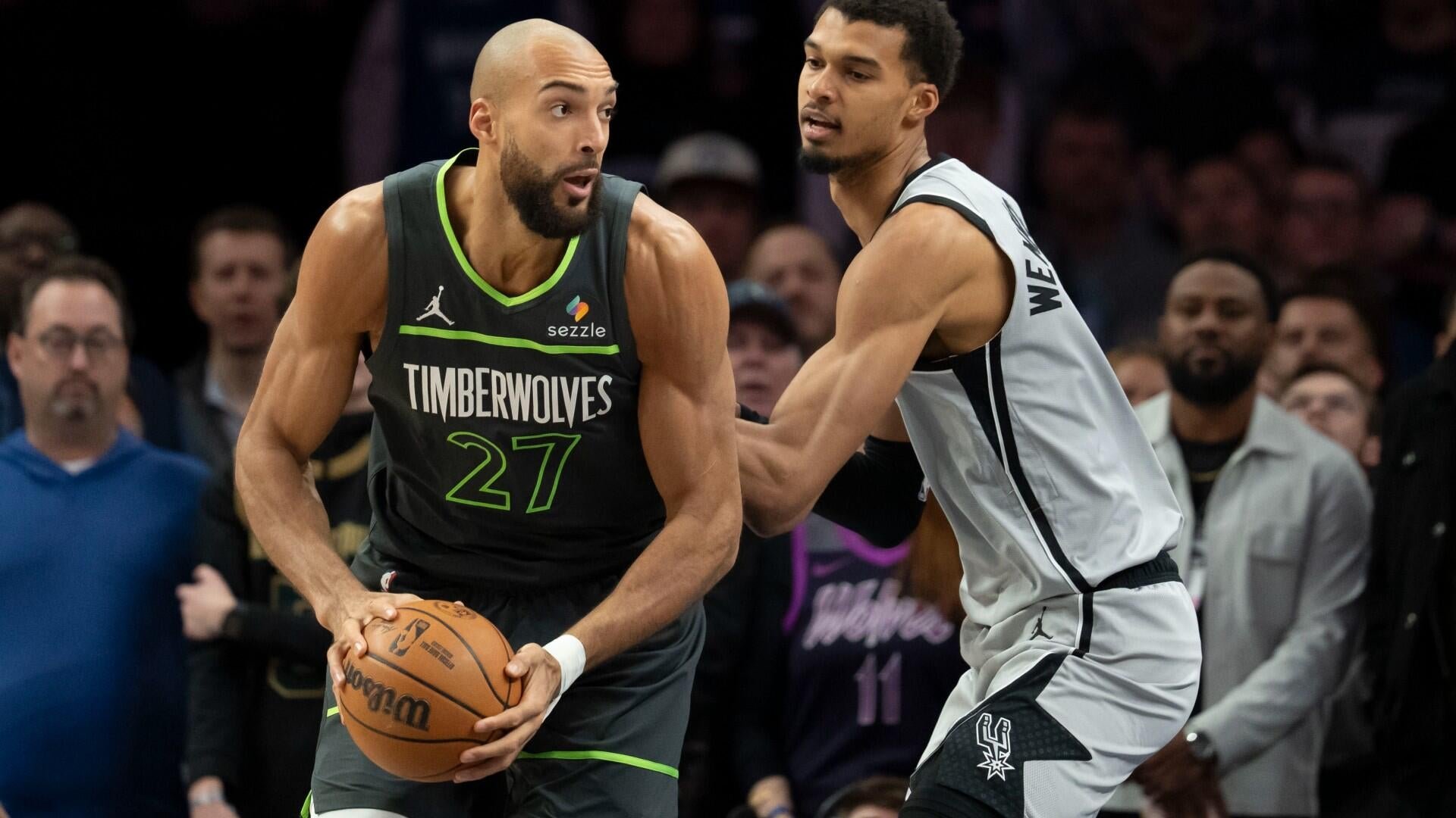 A Timberwolves player wearing jersey number 27 holds a basketball while being defended by a Spurs player in a white jersey, with spectators visible in the background.