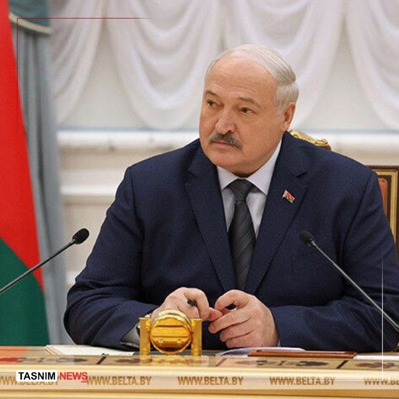 A man in a dark suit with a lapel pin sits at a desk with microphones, holding a pen, with a red-and-green flag visible behind him.