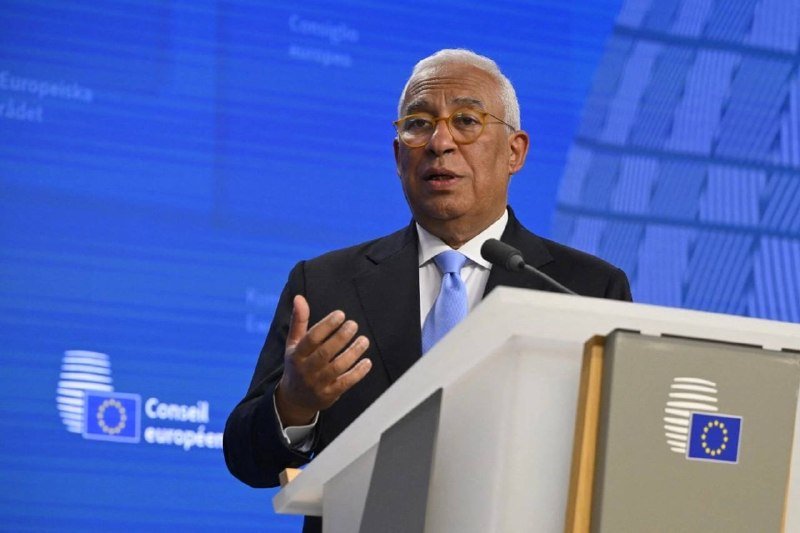 A man in a dark suit and blue tie speaks at a podium bearing the European Council logo, gesturing with his hand against a blue backdrop.