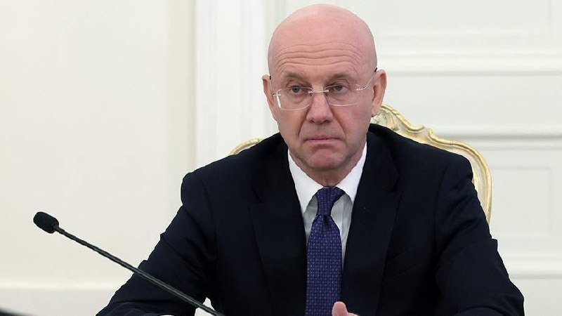 A bald man wearing glasses, a dark suit, and a blue patterned tie sits at a table with a microphone in front of a white paneled wall.