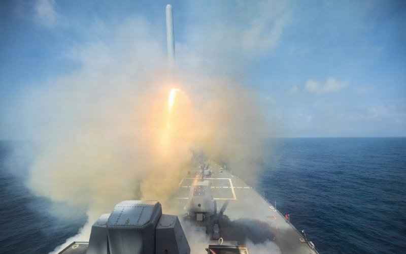 A missile launches vertically from a naval warship's deck, engulfed in bright flame and thick smoke over the open ocean.