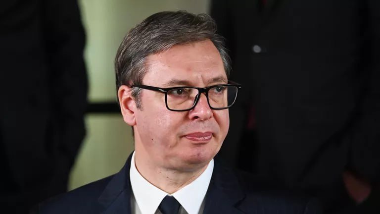 A man with graying hair wearing glasses, a dark suit, white shirt, and tie looks off to the side, with other figures in dark clothing visible in the background.